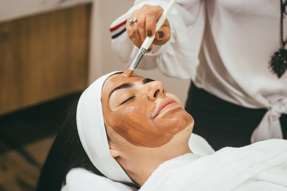 Een masker wordt met een kwast aangebracht op het gezicht van een klant met haarband, in badjas op de behandeltafel