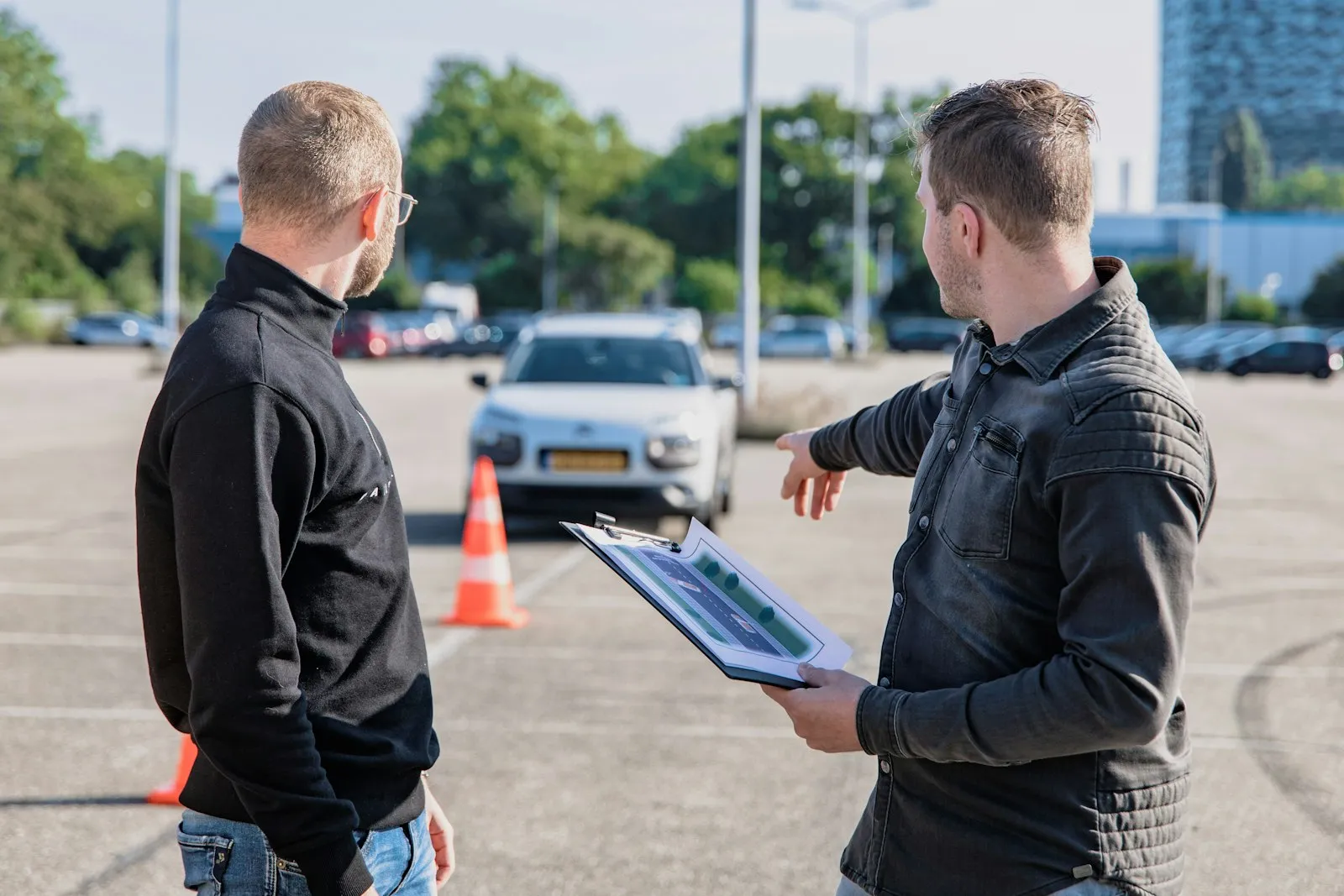 Rij-instructeur bespreekt met een leerling de bijzondere verrichtingen op een oefenterrein, met pionnen en een witte lesauto op de achtergrond