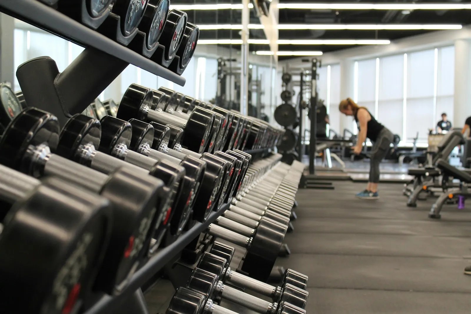 Rijen dumbbells in de lichte krachtzaal van VOLTA, met een sportend lid op de achtergrond