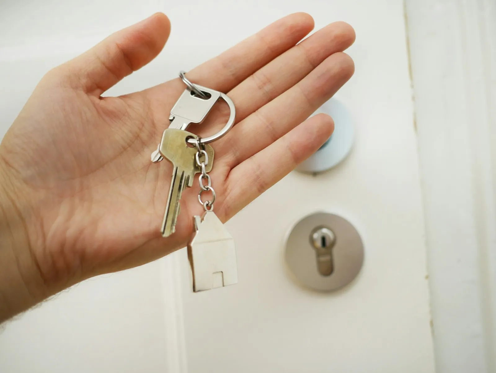 Een hand houdt een sleutelbos met een huisvormige hanger voor een witte voordeur — de sleuteloverdracht