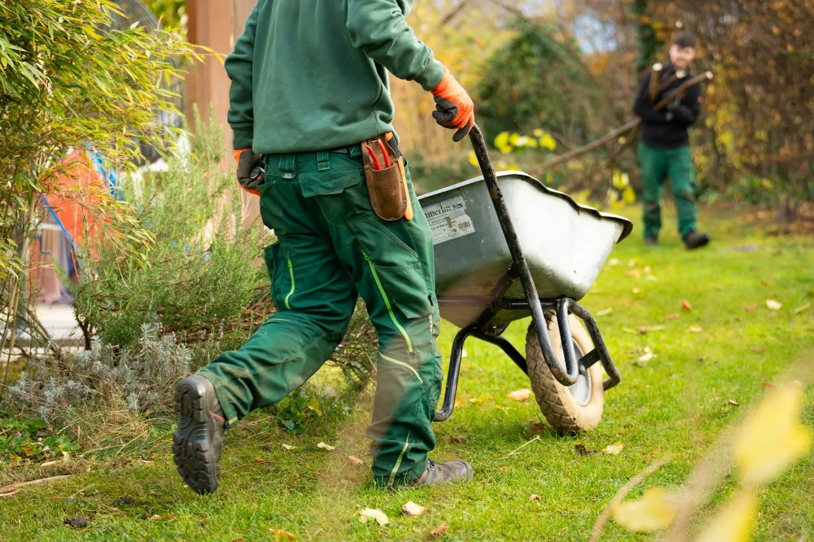 Het team van Grondig Hoveniers aan het werk in een tuin: een hovenier duwt een kruiwagen over het gazon